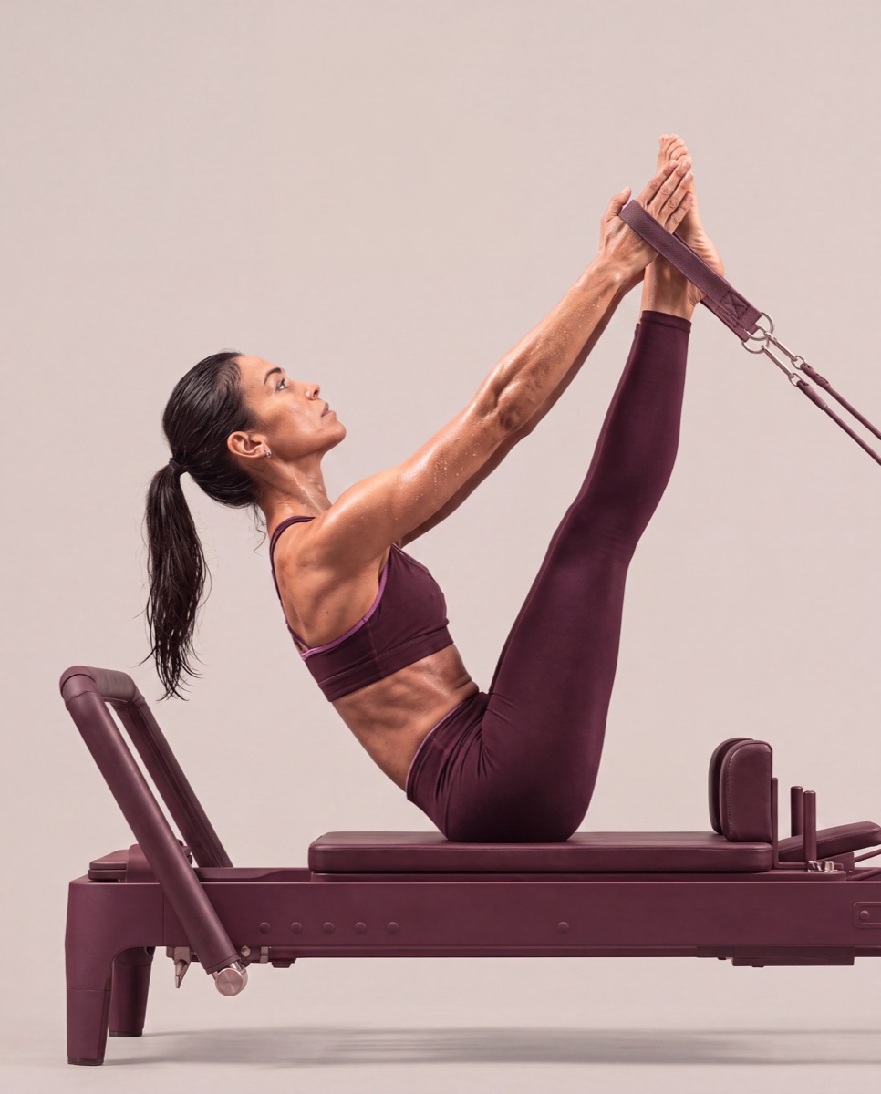 A Like Fred member in a boat-pose teaser on a reformer, in maroon activewear